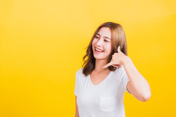 Portrait Asian Thai beautiful happy young woman wear white t-shirt standing smiling white teeth, making call me gesture by finger hand isolated on yellow background, with copy space