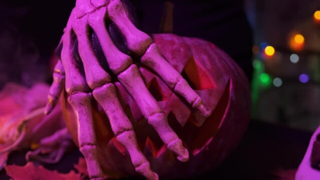 Hand in skeleton costume close-up stroke over the creepy face of carved pumpkin with glowing mouth and eyes against backdrop of thick smoke. Scary cool decoration for Halloween party background.