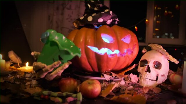 Rotating carved pumpkin in witch hat strokes skull and calls to party Halloween, demanding sweets, trick-or-treat game, among dry leaves, candles, garlands, in dark on doorstep house, close up.
