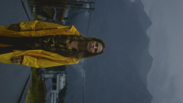 Vertical: Beautiful girl in a yellow raincoat breathing in fresh air. A woman in the mountains enjoys the rain cold foggy autumn day. Travel adventure tourism concept. Road trip wanderlust mood