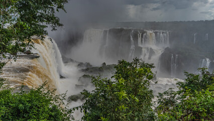 Cascades of waterfalls fall from the ledges of rocks into the river, foaming. Spray and fog rise into the sky. Green vegetation in the foreground. Iguazu Falls. Brazil.