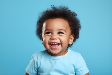 Portrait of a cute African American baby boy wearing blue tee shirt laughing on bright blue background - obrazy, fototapety, plakaty