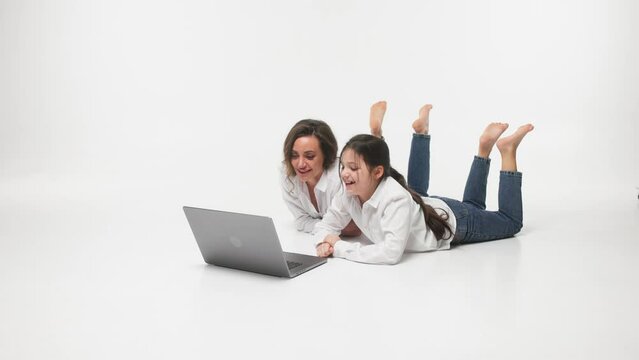 Cheerful smiling happy mother and her little curious daughter lying on the floor and using a laptop to watch cartoons during the weekend