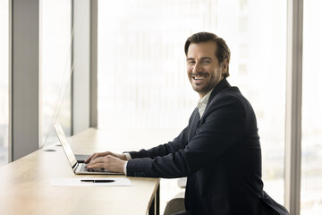 Fototapeta premium Happy business leader man in formal jacket posing at workplace table in office, typing on laptop computer, looking at camera, smiling, posing for professional portrait