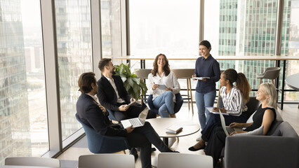 Happy diverse group of business colleagues laughing on meeting in office lobby, modern co-working space, sitting on couch, talking, discussing cooperation, professional tasks