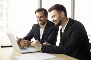 Positive confident business partners men watching content on laptop together, looking at display, laughing, discussing application, software product, online startup project, smiling, laughing