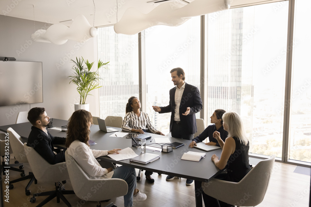 Poster confident handsome adult male boss talking to multiethnic employees on project meeting, giving instr