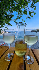 Two glasses of wine and a botte filled with white house wine, placed on a wooden table by the beach in Medveja, Croatia. The table is located in the shadow. Green leaves above. Clear and hot day.