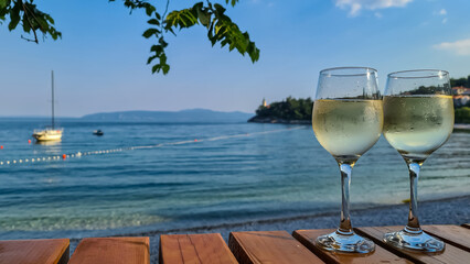 Two glasses of wine filled with white house wine, placed on a wooden table by the beach in Medveja, Croatia. The table is located in the shadow. Green leaves above. Clear and hot day. Summer holiday