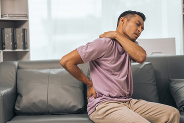 Middle-aged Asian Indian man suffering from back pain, spine and neck pain. sitting on the sofa - Powered by Adobe