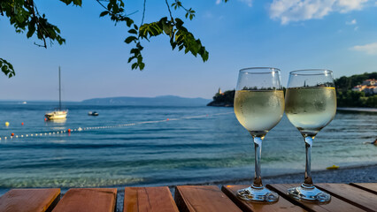 Two glasses of wine filled with white house wine, placed on a wooden table by the beach in Medveja,...