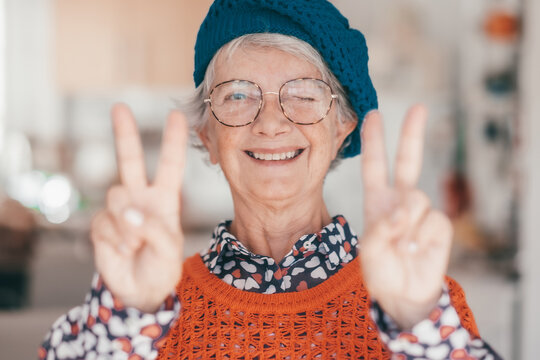 Portrait of happy senior woman wearing glasses and cap looking at camera gesturing positive victory sign with hands. Joyful grandmother winking