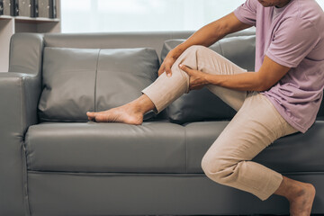 Middle-aged Asian Indian man suffering from calf pain, thigh pain, sitting on the sofa. - obrazy, fototapety, plakaty