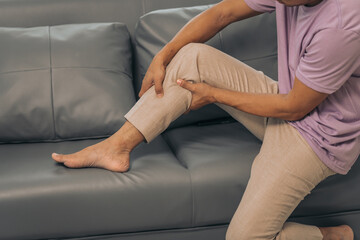Middle-aged Asian Indian man suffering from calf pain, thigh pain, sitting on the sofa.