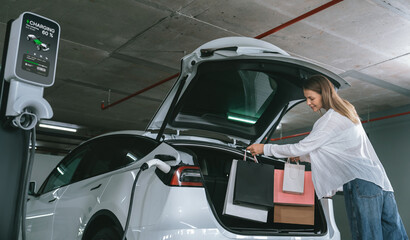 Young woman travel with EV electric car to shopping center parking lot charging in downtown city showing urban sustainability lifestyle by green clean rechargeable energy of electric vehicle innards