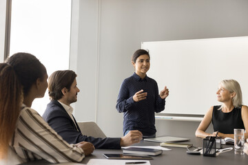 Positive pretty young Indian speaker woman presenting work strategy, teamwork plan, idea for startup project, standing at table on meeting, talking to listening multiethnic coworkers