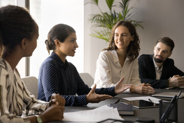 Confident serious young Indian professional girl sharing creative ideas with colleagues on brainstorming meeting, speaking to diverse team of listening coworkers, offering business plan
