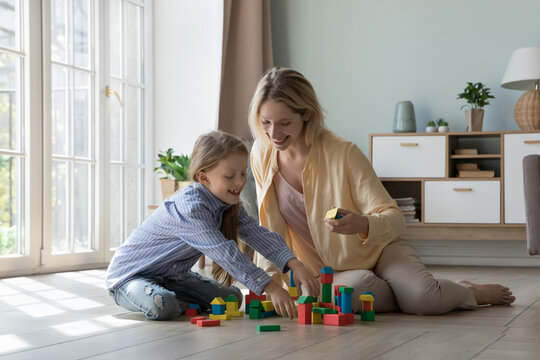 Cheerful excited mom and kid playing learning game at home, constructing toy town model from building blocks on heating floor, enjoying family leisure time, motherhood