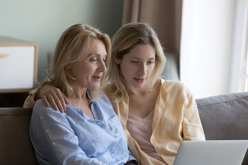 Focused positive mature mother and beautiful young grownup daughter using online ecommerce app on laptop, watching movie, interactive TV channel, enjoying domestic wireless connection, communication