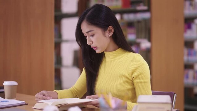 Concentrated young Asian female student in yellow clothes reading books research for study on table in college public library.