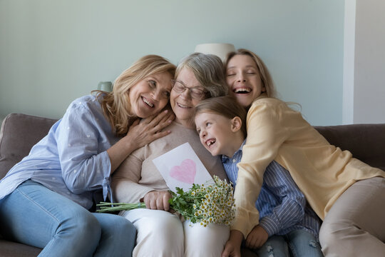 Cheerful girls and women congratulating eldest great grandma on birthday, giving flowers, hand drawn greeting card with heart from kid. Four female family generation hugging, celebrating mothers day
