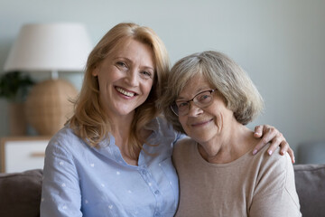 Grownup mature daughter and elderly mother posing at home, looking at camera, smiling, hugging with gratitude, care, love, enjoying family leisure time. Two female generation head shot portrait