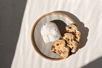Stollen, sweet fruit bread with raisins and sugar powder sliced on pieces on neutral beige table background with aesthetic sunlight shadow. Delicious morning breakfast.