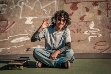 teenager man with headphones and skateboard sitting at skate park doing ok sign with finger