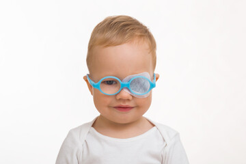 1 year and 4 month old cute smiling baby boy with blue eyeglasses and white sticking plaster isolated on light gray background. Front view. Closeup. Looking at camera. Amblyopia or lazy eye treatment.