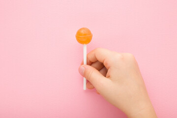 Little child fingers holding orange lollipop on stick on light pink table background. Pastel color. Sweet candy. Closeup. Top down view. - obrazy, fototapety, plakaty