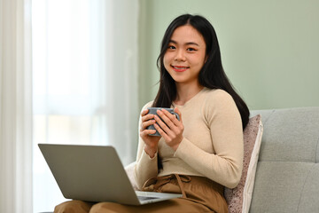 Portrait of pretty young woman sitting on sofa with laptop and drinking coffee. People, technology and lifestyle