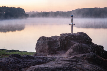 l'épée légendaire Excalibur plantée sur son rocher devant un étang dans la brume du matin au lever du soleil - obrazy, fototapety, plakaty