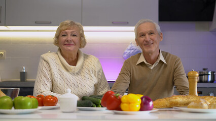 Portrait of smiling senior spouses sitting in the kitchen, enjoying a healthy lunch with vegetables
