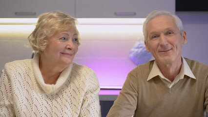 Fototapeta na wymiar Loving mature wife looking at her smiling handsome senior husband sitting in the kitchen