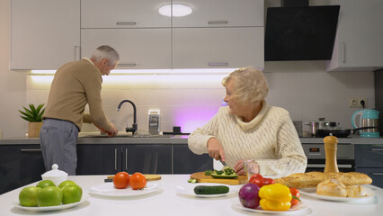 A senior woman watches her husband washing fresh vegetables from their garden while cooking in the kitchen