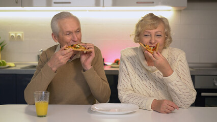 Senior man and woman eating pizza in the kitchen, enjoying take-out, fast food