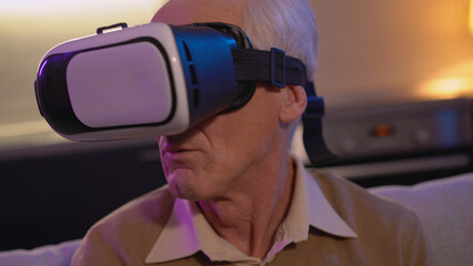 A senior man is sitting on the sofa, wearing a virtual reality headset, immersed in cyberspace in a close-up shot
