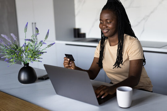 African Woman Using Credit Card While Sitting At Desk With Laptop Paying Bills In Internet Or Transferring Money Via App