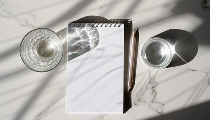 Aesthetic white morning flat lay. Glass with water, blank notebook on marble table background. Natural dappled light shadows. Minimal business template, copy space