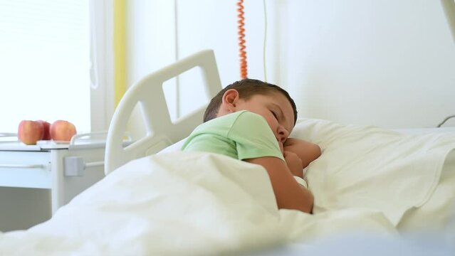A caring young mother covers her little son with a blanket and kisses him as he lies in a hospital bed in a hospital room.