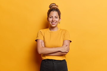 Horizontal shot of lovely European woman with hair bun keeps arms folded stands satisfied poses for making photo against vivid yellow background dressed in casual t shirt and black trousers. - 658585951