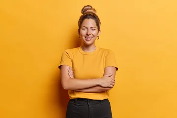 Foto op Canvas Horizontal shot of lovely European woman with hair bun keeps arms folded stands satisfied poses for making photo against vivid yellow background dressed in casual t shirt and black trousers. © Wayhome Studio