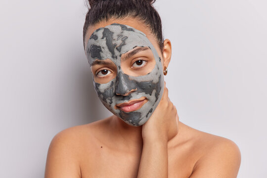 Beauty concept. Indoor close up of young happy pretty European female applying clay face mask to make skin smooth and fresh standing in centre isolated on white background looking straight at camera