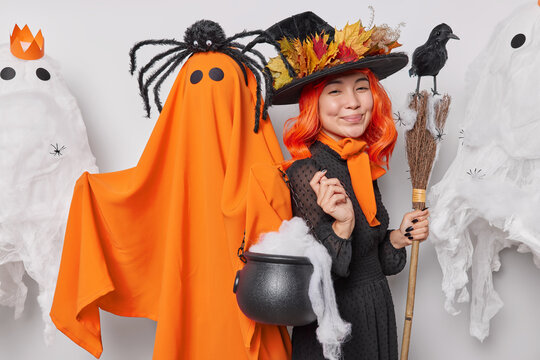 Happy pleased woman with cauldron and broom stands near orange ghost wishes you happy Halloween poses near sppoky creatures preapres for masquerade isolated over white background. 31st October