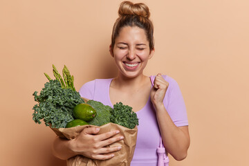 Happy European woman with hair bun clenches fist carries paper bag full of green vegetables like asparagus and avocado rejoices good news sport results has aim to loose weight poses with skipping rope