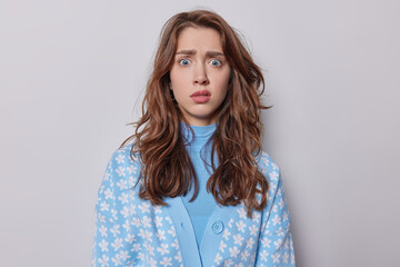 Studio waist up of young beautiful upset European lady with long wavy brunette hair standing isolated in centre on white background looking straight at camera wearing blue cardigan in white flowers