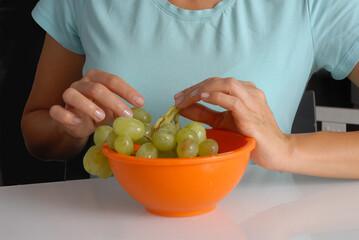 Woman hand holding green grapes bunch on bowl on kitchen table background.