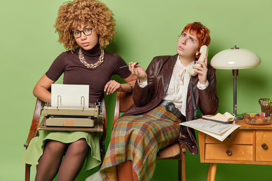 People at work concept. Studio photo of two young concentrated African american and European females sitting in old room on green background typewriting speaking on phone holding pen. Retro style