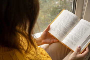 Young beautiful woman sitting by the window yellow knitted sweater read book, daily planner,...