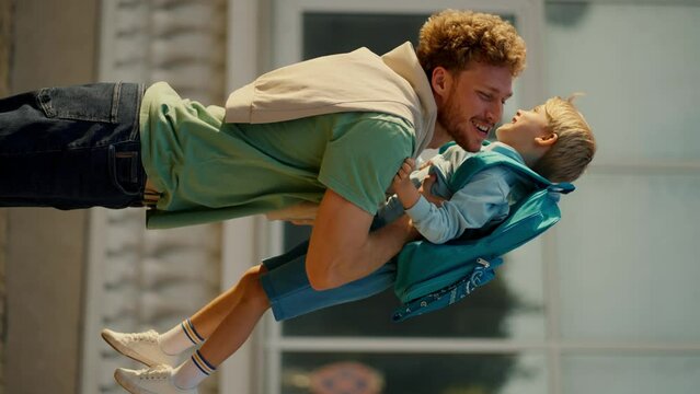 Vertical video A little boy in blue clothes with a blue backpack runs after school to his father in a Green T-shirt with curly hair. Happy meeting of father parent and his little boy after school